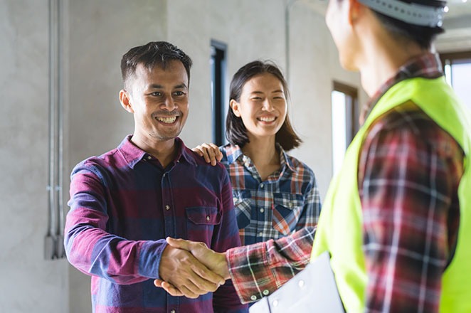 AA contractor in a safety hat and vest shakes the hand of a smiling homeowner, his wide is behind him also smiling